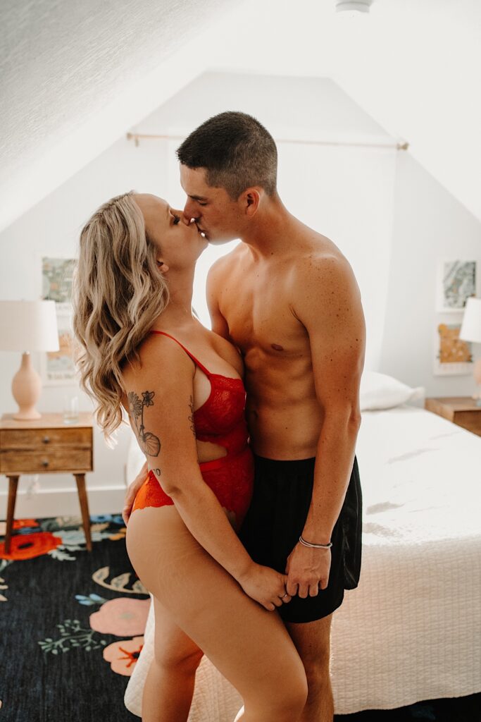A woman in red lingerie kisses a man in his underwear while they stand next to a bed