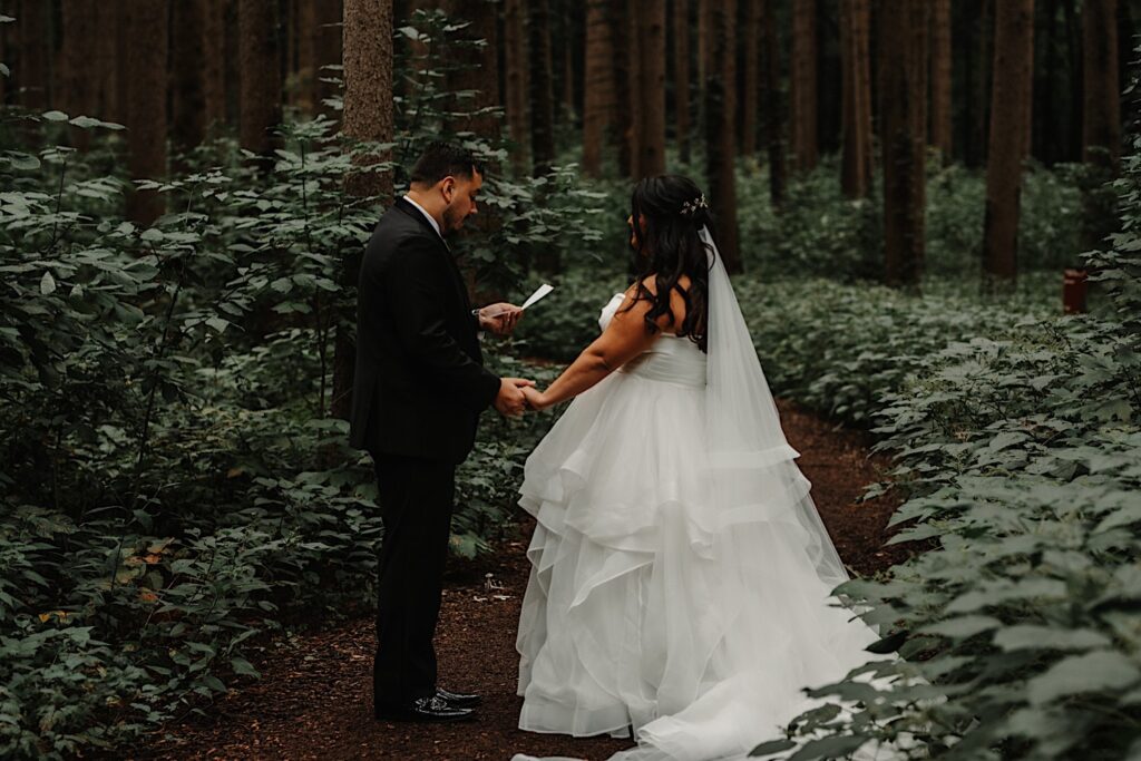 A bride and groom hold hands while in a forest near their wedding venue, the groom is reading his vows to the bride