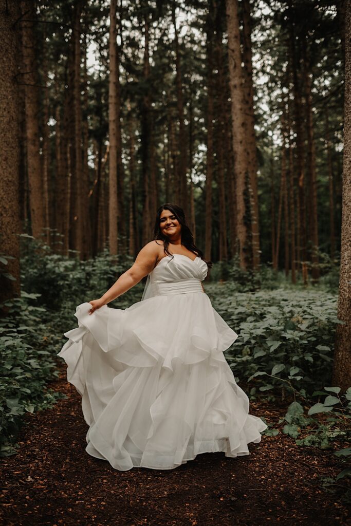 A bride stands in a forest and spins in her dress while smiling at the camera