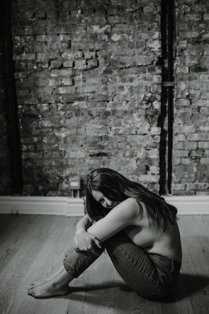 Black and white photo of a woman wearing only jeans sitting on the floor of a brick building and smiling at the camera during a boudoir session