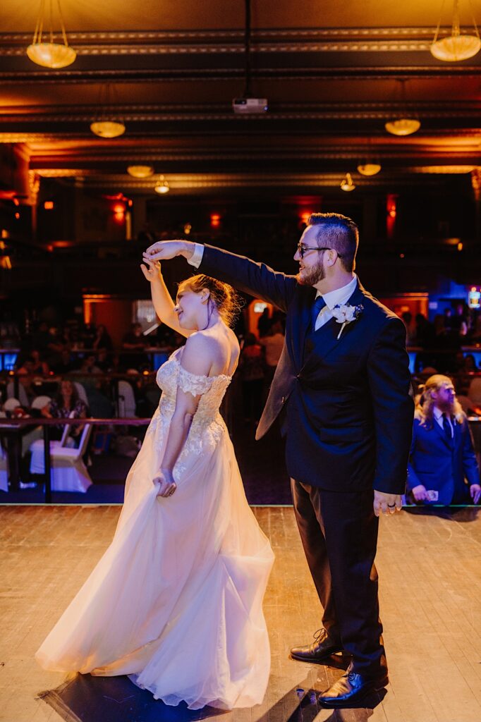 A bride and groom dance together while on stage as guests of their reception watch in the background