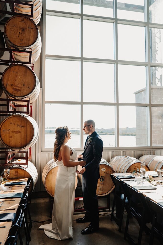 A bride and groom hold hands while standing in front of a window and whiskey barrels