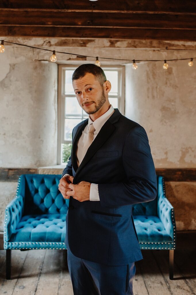 A groom looks at the camera while standing in a room in front of a blue couch as he buttons his suit coat