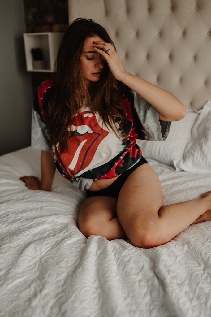 A woman in a Rolling Stones t shirt and panties sits on a bed and plays with her hair during a boudoir session