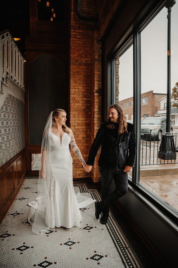 A bride and groom hold hands and smile at one another in a brick building as the groom leans on a large window