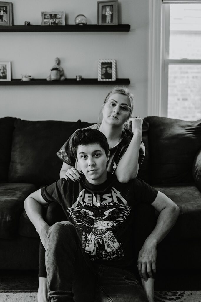 Black and white photo of a couple looking at the camera, the woman is sitting on the couch behind the man who is sitting on the floor