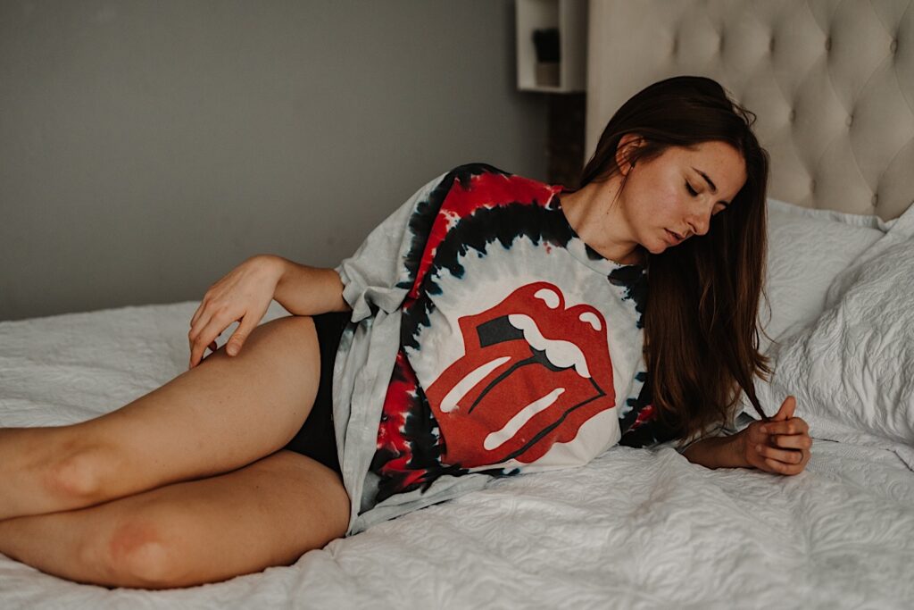 A woman in a Rolling Stones t shirt and panties lays on a bed and plays with her hair while taking boudoir photos in the suburbs of Chicago