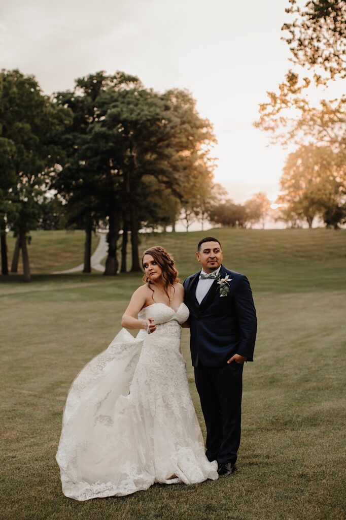 A bride and groom stand side by side in the middle of a field and lookin opposite directions as the sun sets behind them