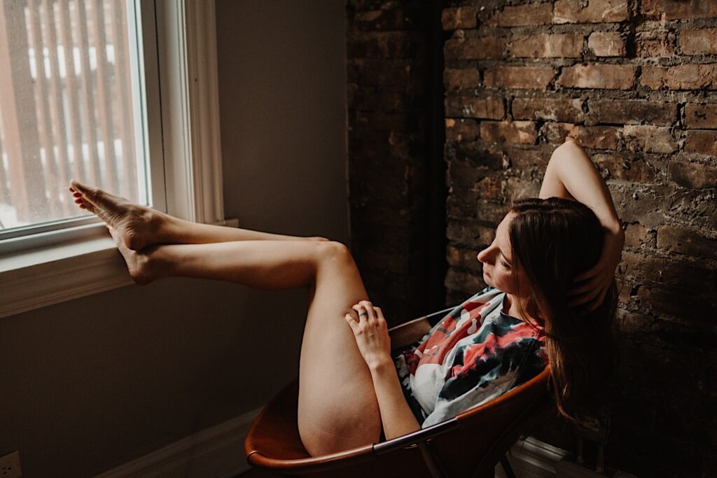 A woman wearing a t shirt and panties sits in a chair and rests her feet on a windowsill while taking boudoir photos in the suburbs of Chicago