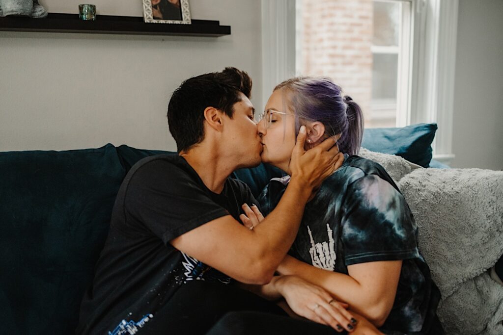 A couple kiss one another while sitting on a couch in their Chicago apartment while taking their engagement photos
