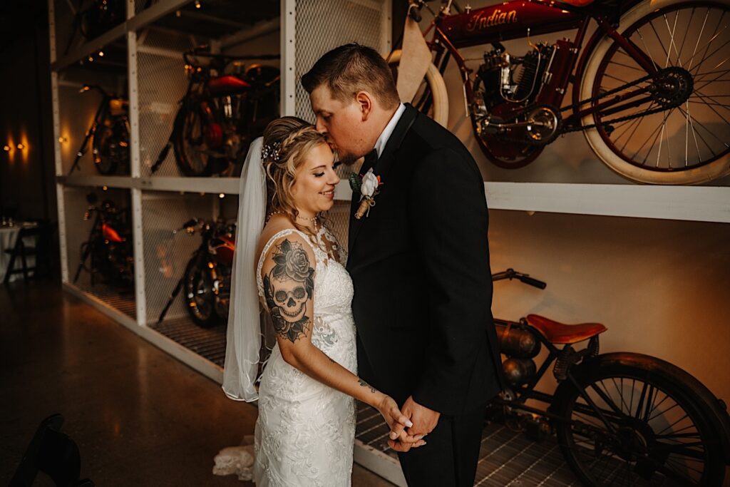 A bride smiles as the groom kisses her on the head while they stand in front of a wall full of classic motorcycles, photo taken by a Central Illinois Wedding Photographer