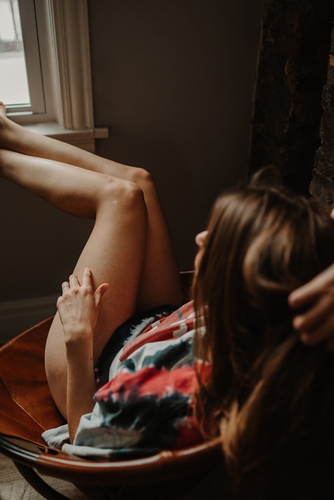 A woman in a t shirt and panties sits in a chair and rests her feet on the windowsill during a boudoir session