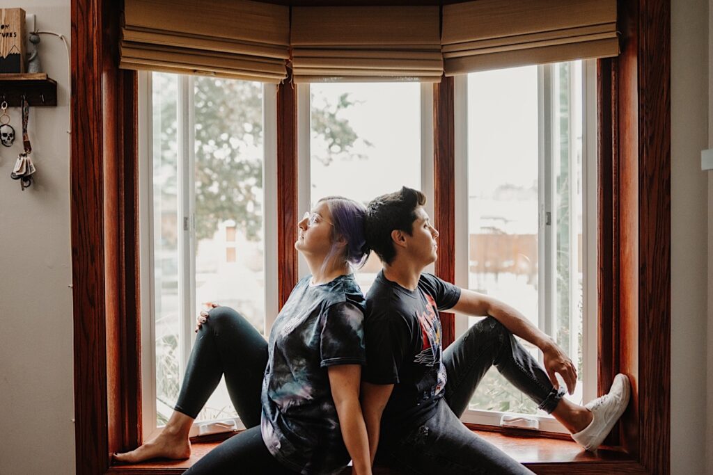 A couple sit back to back on a windowsill in their Chicago apartment while taking engagement photos