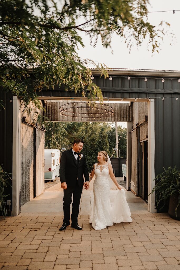 A bride and groom stand side by side and hold hands while outside of their wedding venue 