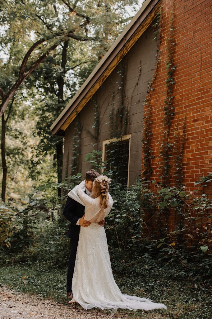 A bride and groom embrace one another while standing outside of a brick house covered in vines