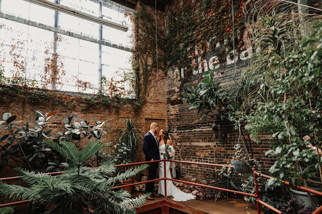 A bride and groom stand side by side inside their wedding venue, behind them are brick walls covered in vines and other plants