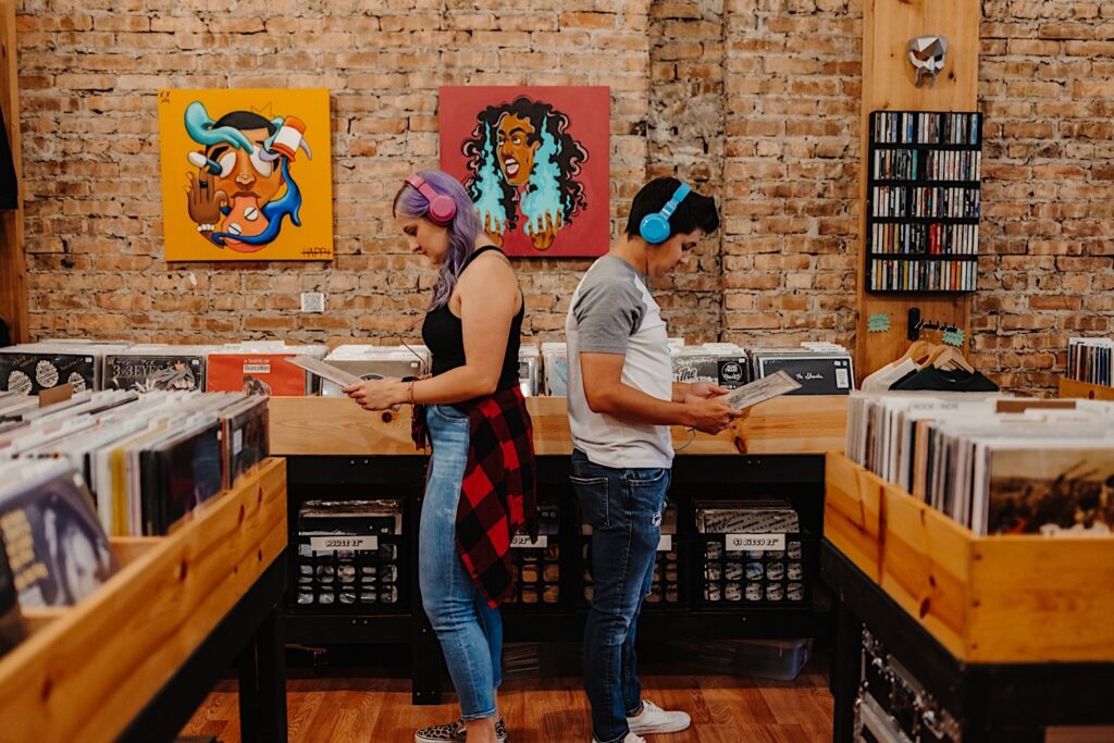 A couple wearing colorful headphones face back to back while looking at records in a Chicago record shop while taking engagement photos