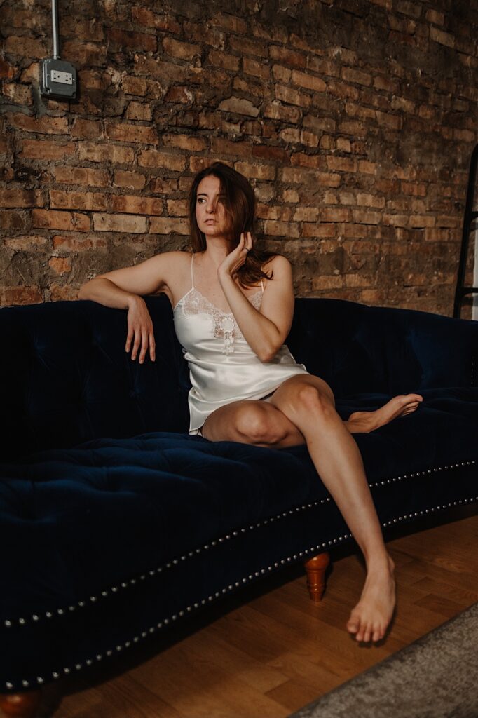 A woman in a white slip dress sits on a blue couch in a brick building while taking boudoir  photos
