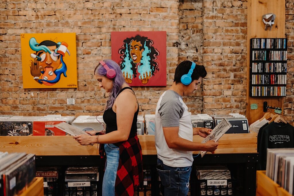 A couple wearing colorful headphones face back to back while looking at records in a Chicago record shop while taking engagement photos