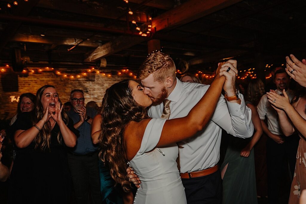 A bride and groom kiss while dancing during their indoor wedding reception as guests around them cheer, photo taken by a Central Illinois Wedding Photographer