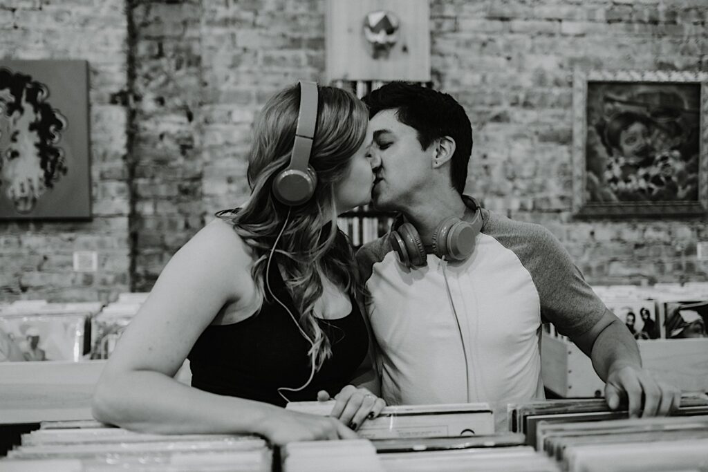 Black and white photo of a couple kissing one another while looking through records in a vinyl shop in Chicago while taking engagement photos