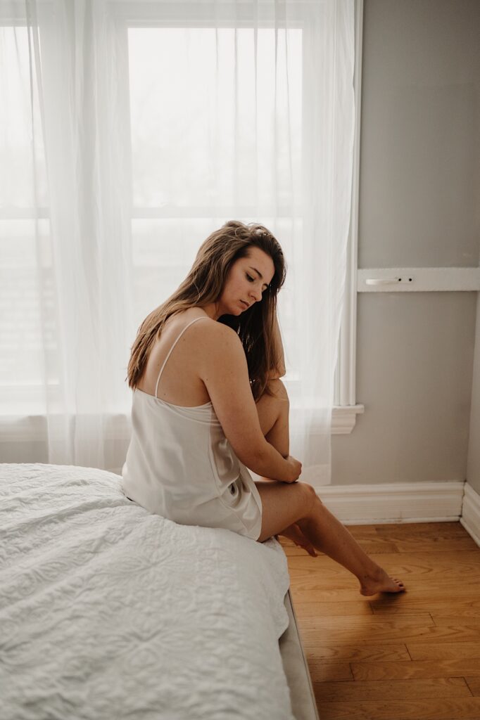 A woman in a white slip dress sits on a bed and plays with her hair during a boudoir session