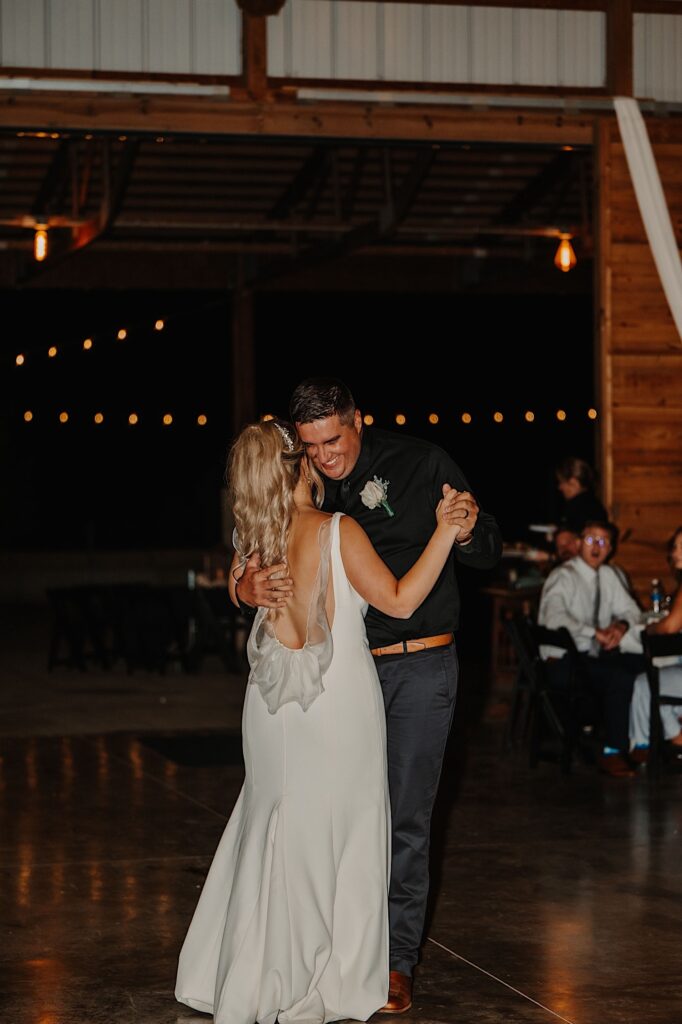 A groom smiles as he and the bride share their first dance during their indoor wedding reception