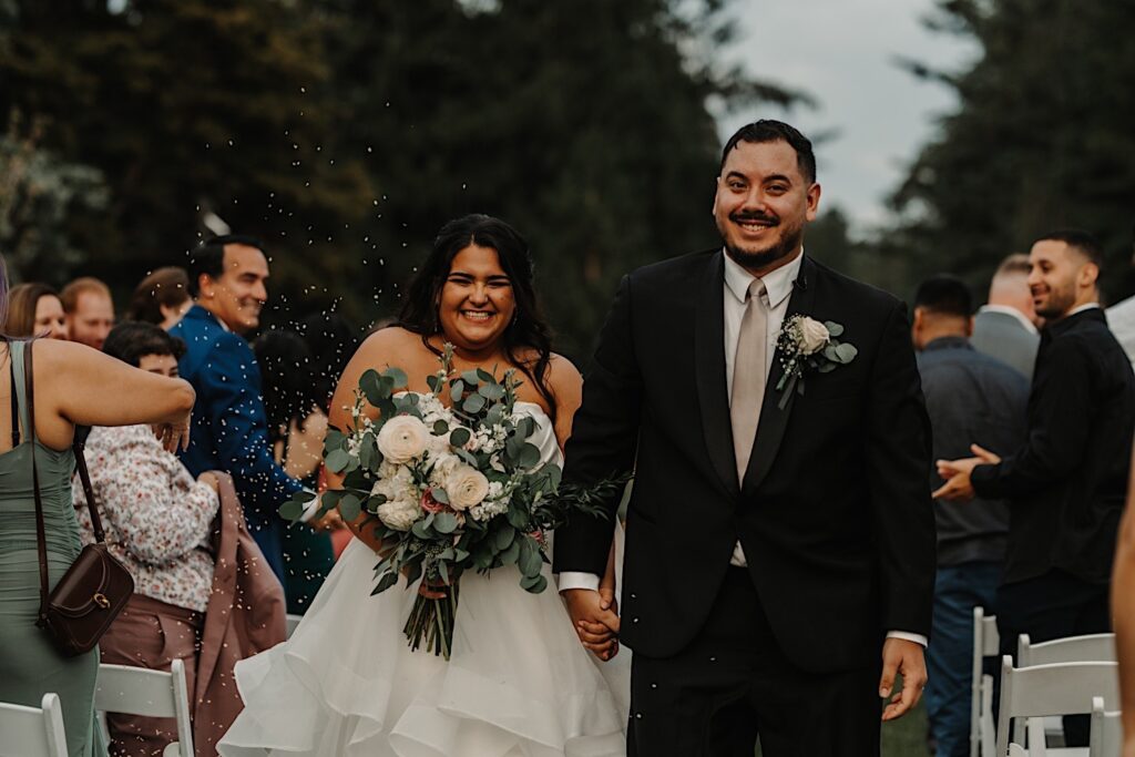 A bride and groom smile at the camera while walking hand in hand down the aisle as guests throw rice in the air, photo taken by a Central Illinois Wedding Photographer