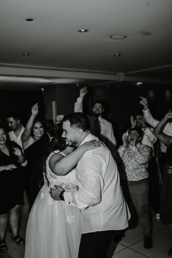 Black and white photo of a bride and groom dancing together during their indoor wedding reception as guests cheer behind them