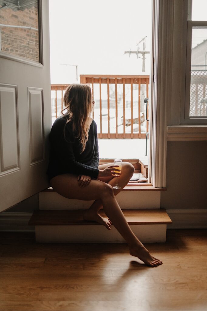 A woman in a sweater and panties sits in an open doorway and looks out at the snow while holding a glass of wine during a boudoir session