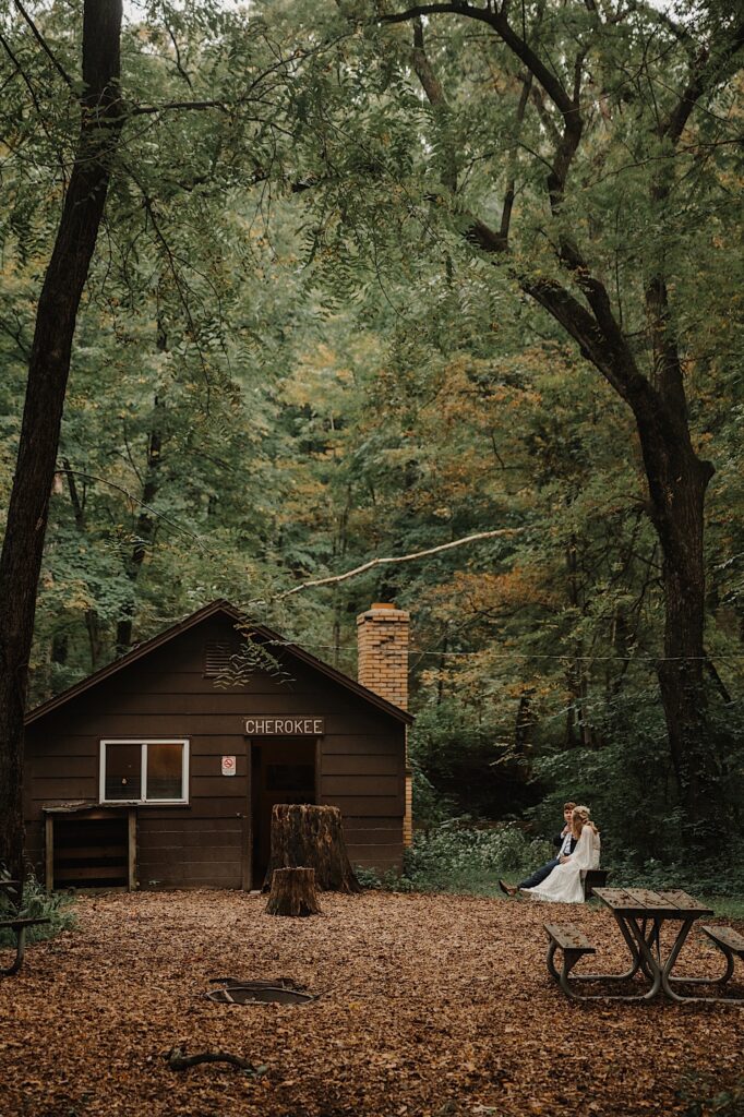 A bride and groom sit next to one another on a bench next to an old cabin in the woods