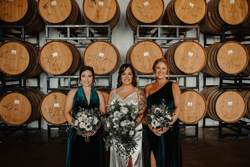 A bride and two of her bridesmaids stand together and smile at the camera in front of a wall of bourbon barrels, photo taken by a Central Illinois Wedding Photographer