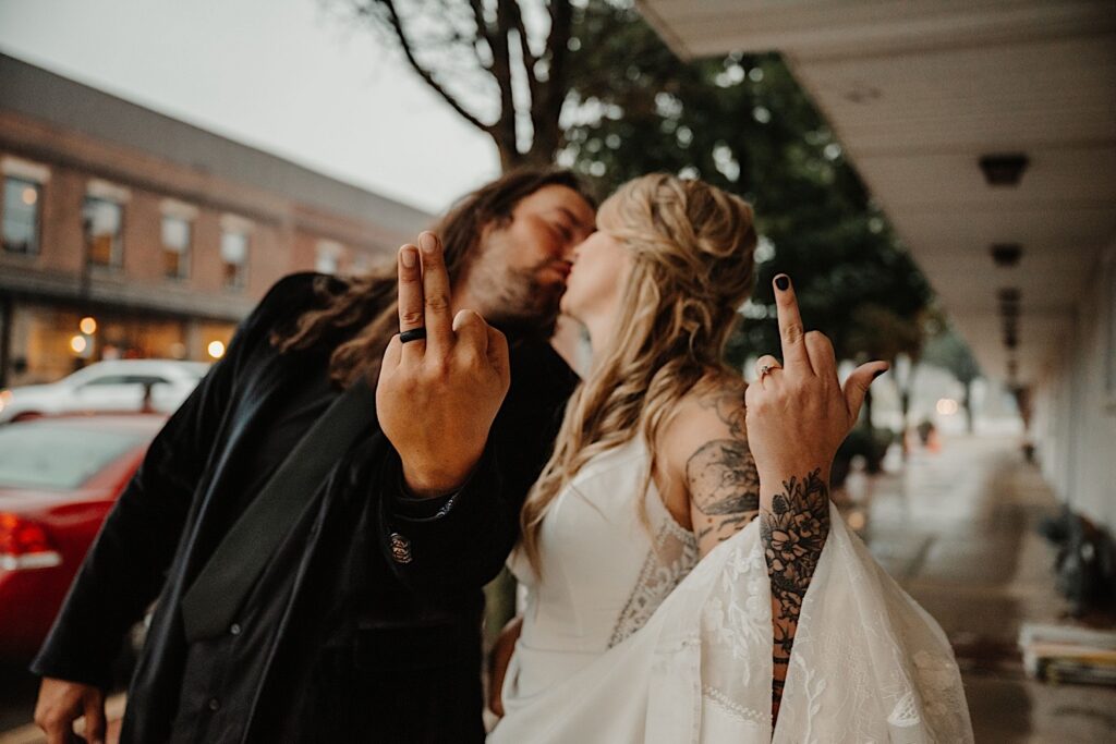 A bride and groom kiss one another while standing on a sidewalk, the groom has his ring and middle finger up to the camera while the bride only has her middle finger up, photo taken by a Central Illinois Wedding Photographer