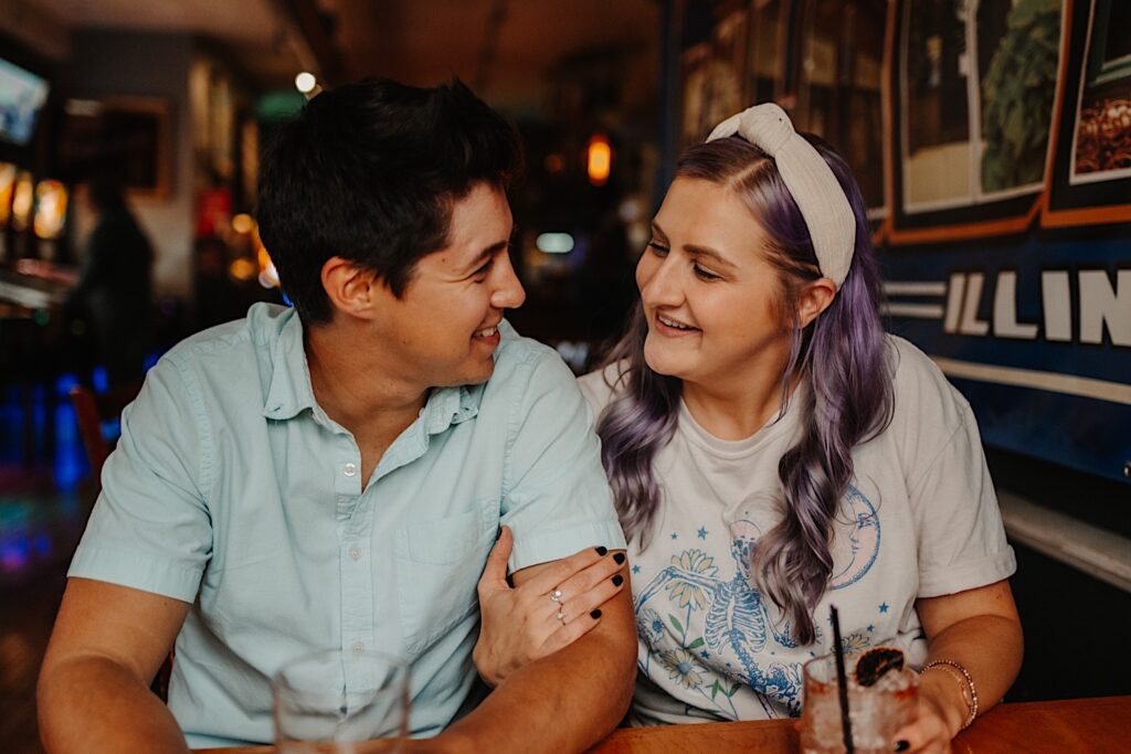 A couple smile at one another while sitting together at a table in a Chicago bar and have their engagement photos taken