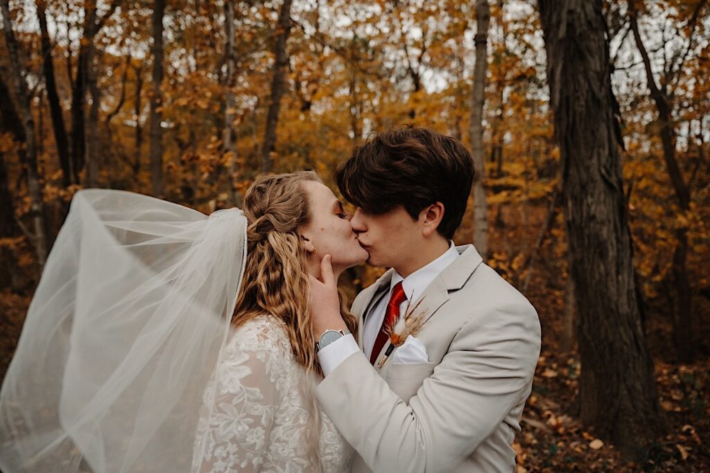 A bride and groom kiss one another while standing in a forest with fall colored leaves behind them, photo taken by a Central Illinois Wedding Photographer