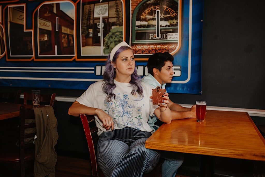 A couple sit together at a table in a bar of Chicago, the woman is holding her drink while the man has his on the table as they have their engagement photos taken