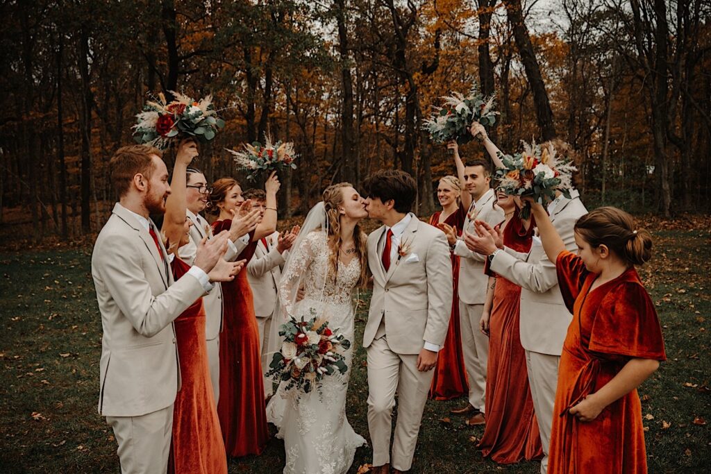 A bride and groom kiss one another while members of their wedding party cheer on either side of them, they are outdoors and behind them is a forest with fall colored leaves all around, photo taken by a Central Illinois Wedding Photographer