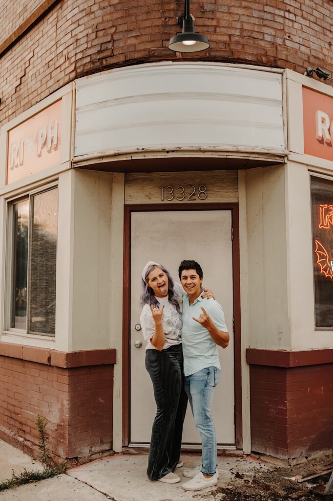 A couple stand outside of a bar in Chicago and embrace while making the "rock and roll horns" gesture towards the camera and smiling