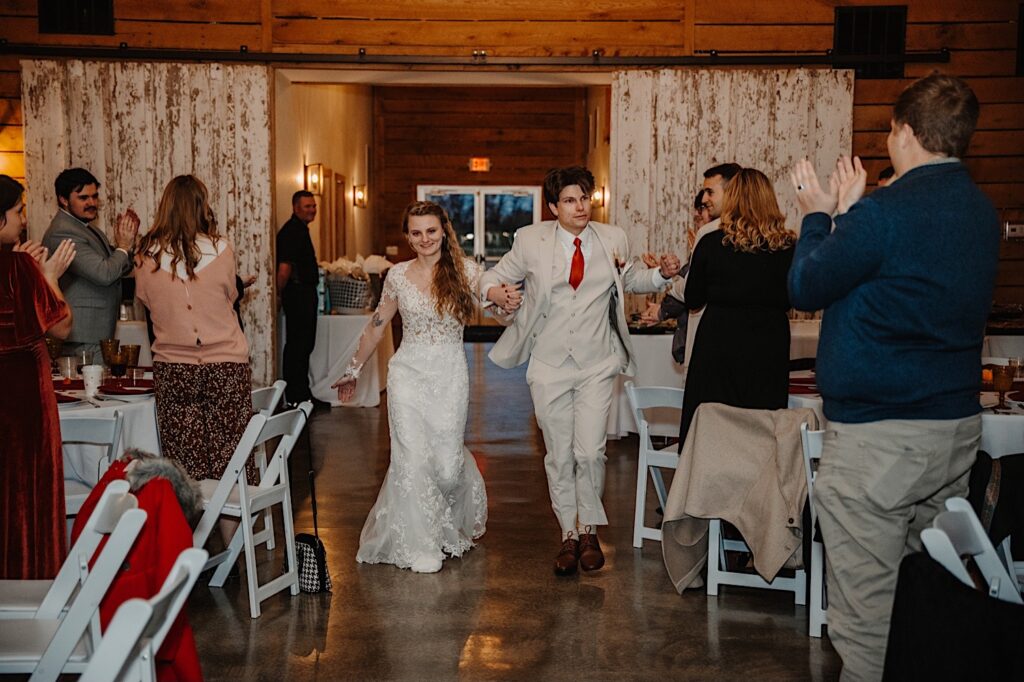 A bride and groom hold hands as they enter their indoor wedding reception as guests stand and cheer around them, photo taken by a Central Illinois Wedding Photographer