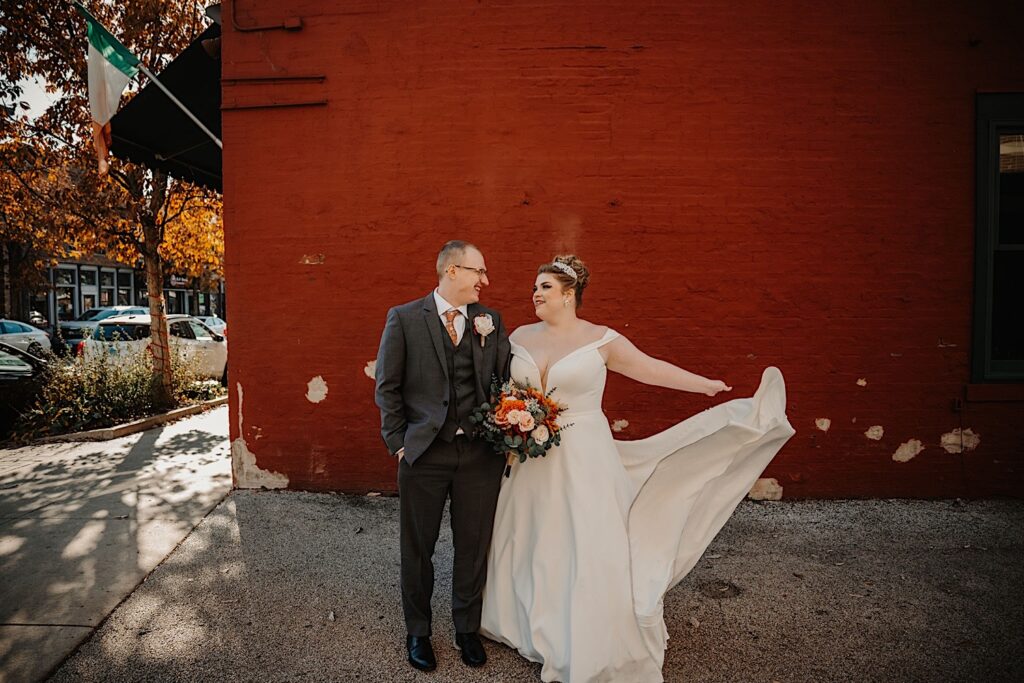 A bride and groom stand next to one another and smile as the bride plays with her dress while they stand in front of a red brick wall, photo taken by a Central Illinois Wedding Photographer