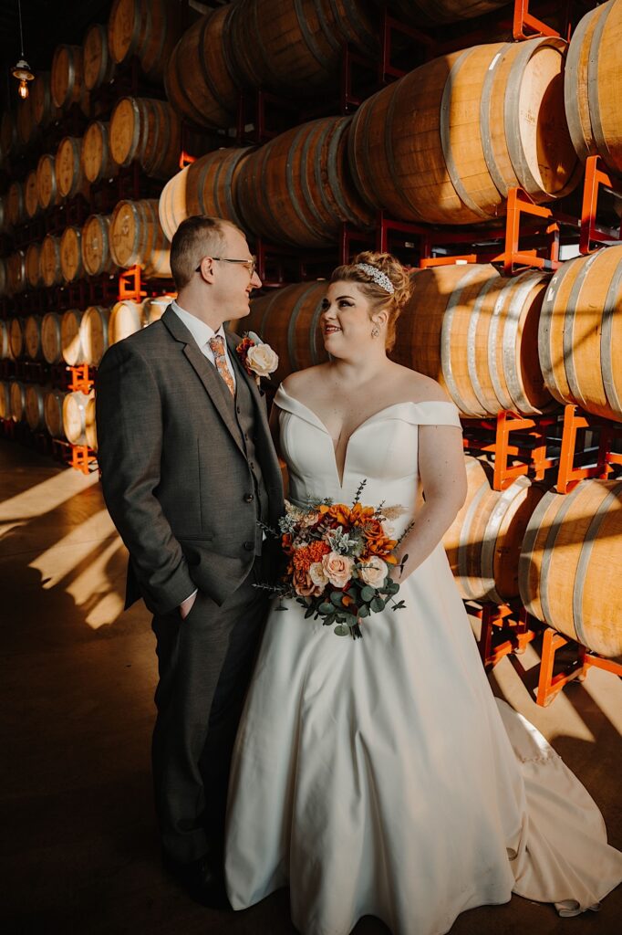 A bride and groom stand side by side and smile at one another while standing in front of a wall of bourbon barrels