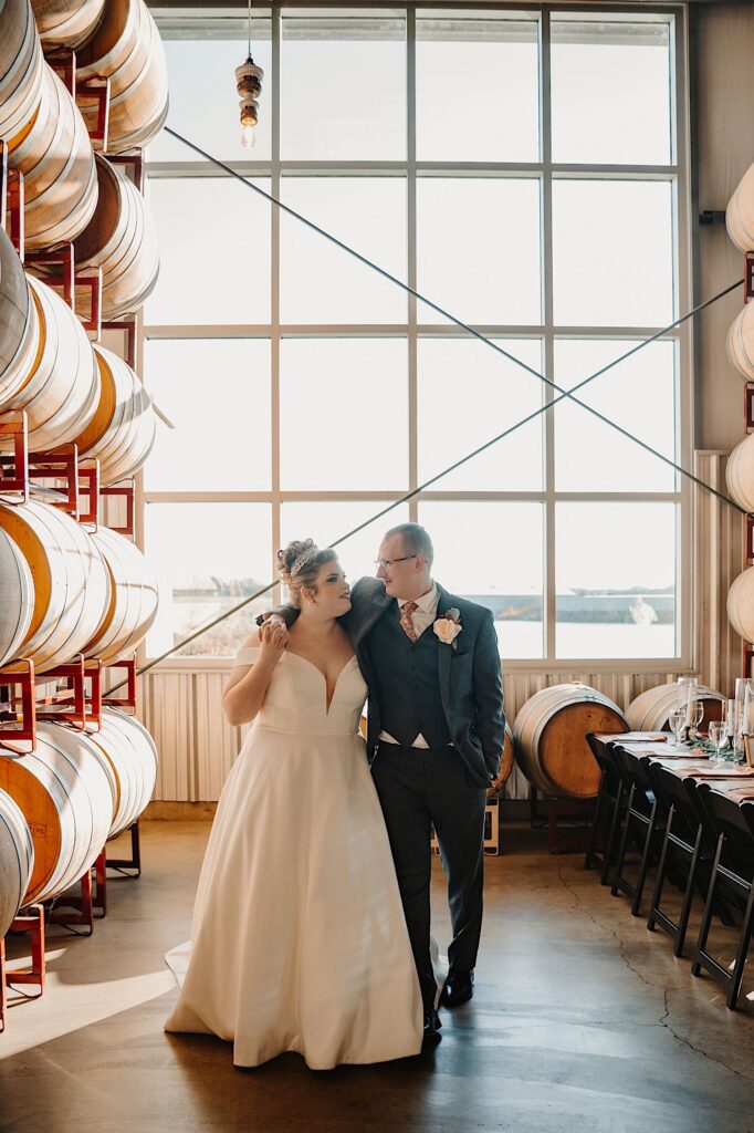 A groom wraps his arm around the bride as they smile at one another, on the wall besides them are stacked bourbon barrels