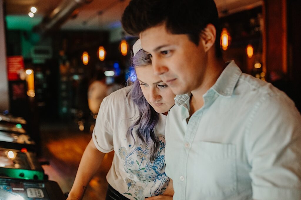 A woman leans on a man's shoulder while he plays pinball in a bar of Chicago while having their engagement photos taken