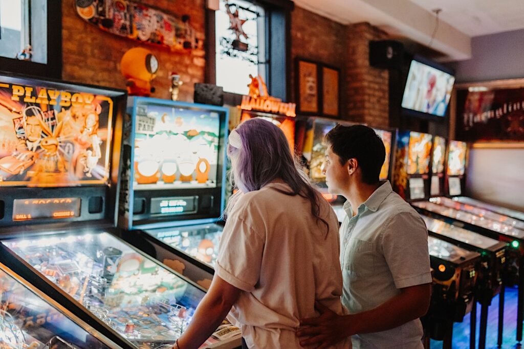 A man puts his arm around a woman while she plays pinball in a bar of Chicago while their engagement photos are taken