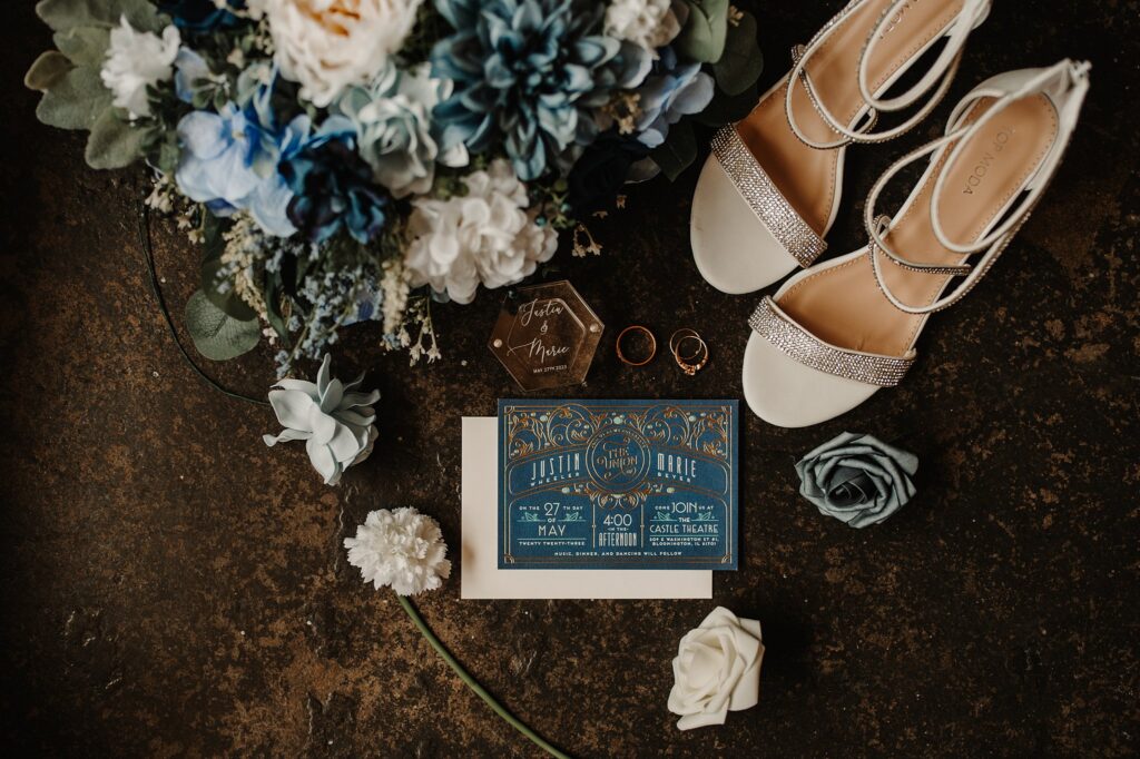 wedding detail photo of wedding invitation, shoes, florals, and rings against concrete floor at the castle theater in bloomington illinois